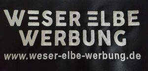 Logo Weser Elbe Werbung - Firmendruck auf schwarzer Fleecejacke Weser Elbe Werbung Stickerei in Osterholz-Scharmbeck | Textilveredelung, Stickarbeiten und Folienaufdruck in OHZ